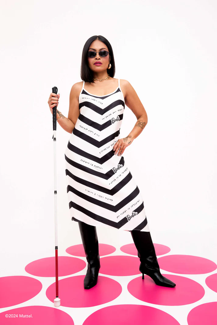 Woman with walking stick and sunglasses wears black and white chevron dress and black boots, standing on pink dotted floor.  Phrases in braille say "original style icon," "changing the world," "inspiring since 1959," "limitless possibilities," "powered by purpose," "fashion is for everyone," and "the future is inclusive."